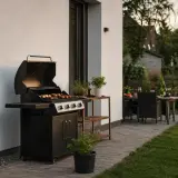 Nowoczesny grill w ogrodzie, gotowy na letnie przyjęcie. Na ruszcie szaszłyki, obok stolik z ziołami i doniczką. W tle stół z krzesłami i zielony trawnik.
