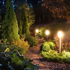 Nastrojowe oświetlenie rabaty tworzy magiczną atmosferę. Ścieżka z kamieni i drewna prowadzi przez bujną zieleń, podkreśloną przez nowoczesne lampy ogrodowe.