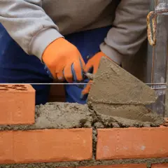 Murarz w pomarańczowych rękawicach układa cegły, tworząc solidny grill z cegły.