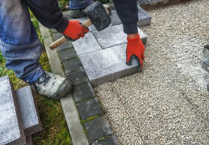 Ręce w czerwonych rękawiczkach układają kostkę granitową, tworząc solidny chodnik. Młotek czeka na użycie.