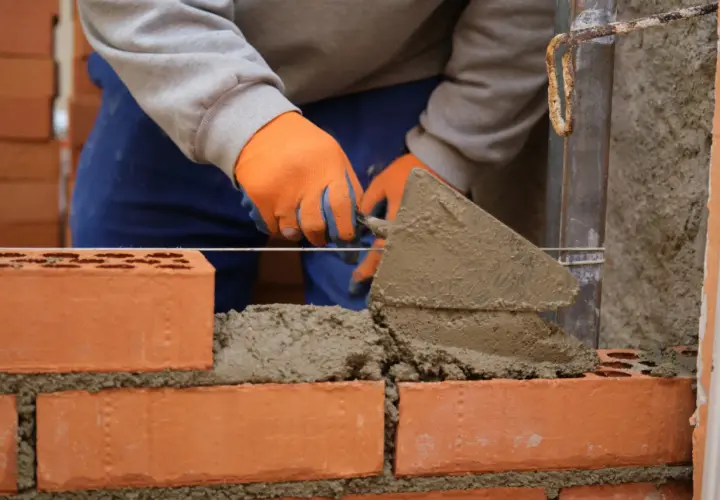Murarz w pomarańczowych rękawicach układa cegły, tworząc solidny grill z cegły.
