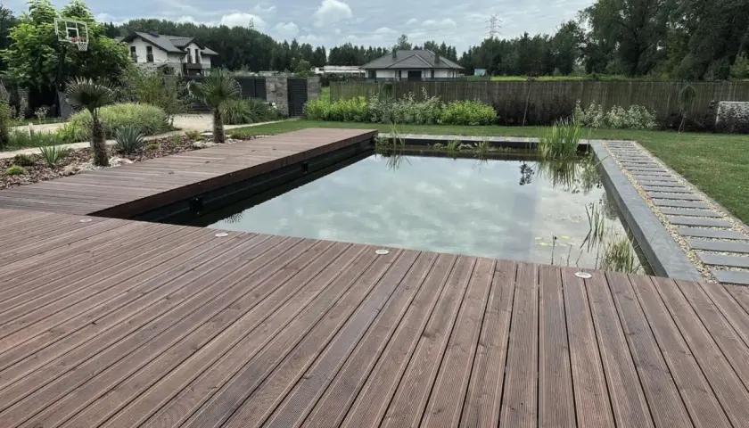 Drewniany taras otacza nowoczesny staw kąpielowy z roślinnością. Idealne miejsce na relaks w ogrodzie.