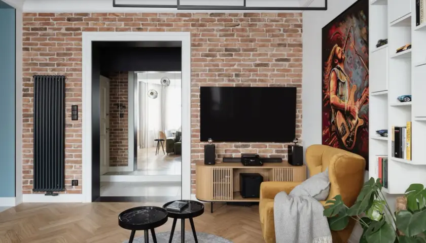 Nowoczesny salon z ceglaną ścianą, na której wisi telewizor. Oświetlenie podkreśla fakturę cegły, tworząc przytulną atmosferę.