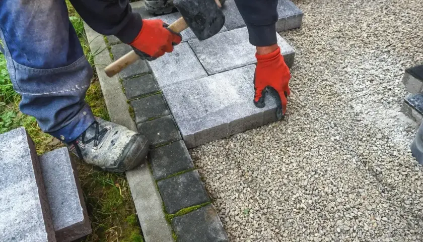 Ręce w czerwonych rękawiczkach układają kostkę granitową, tworząc solidny chodnik. Młotek czeka na użycie.
