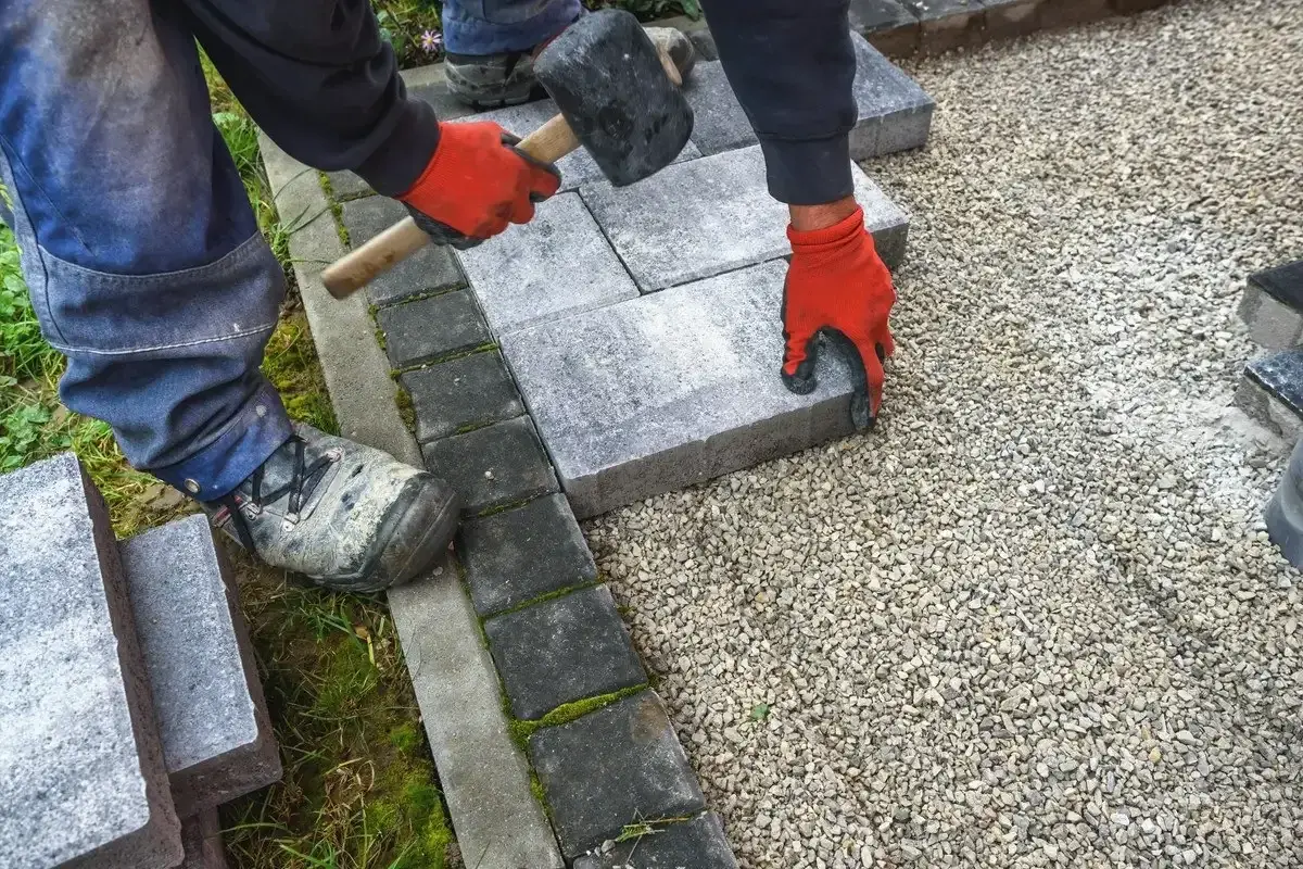 Ręce w czerwonych rękawiczkach układają kostkę granitową, tworząc solidny chodnik. Młotek czeka na użycie.