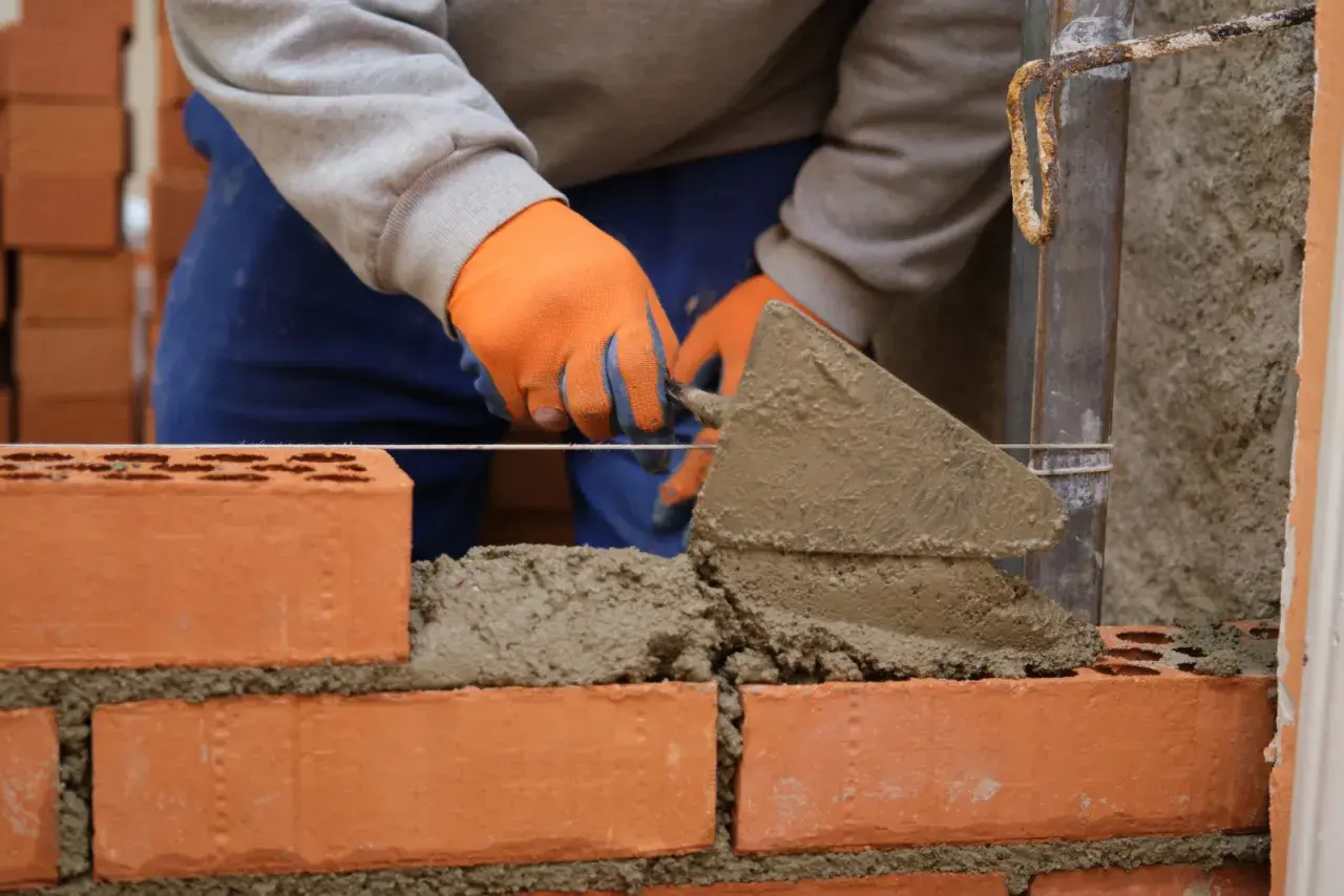 Murarz w pomarańczowych rękawicach układa cegły, tworząc solidny grill z cegły.